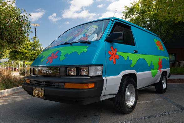 A Japanese panel van, colored blue with the bright designs of the Scooby Doo van. A sun reflector with a picture of the Scooby Doo cast is placed behind the windshield.