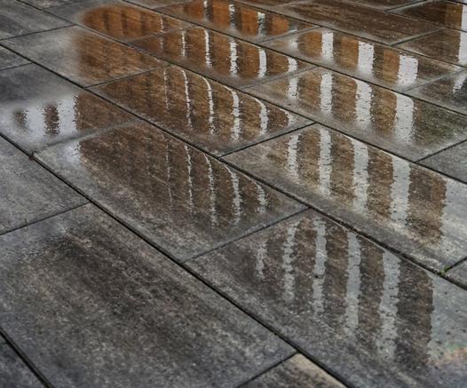 A low-angle close-up shot of wet, dark grey rectangular paving slabs on the ground. The surface is slick with water, creating strong reflections of vertical elements, likely railings or fence posts, appearing as shimmering white and dark brown stripes across the wet flagstones. The paving stones are laid in a stretcher bond pattern, slightly offset from one another. The texture of the wet stone is visible, showing varying shades of dark grey and subtle hints of brown. The reflections distort and stretch the vertical lines, giving the impression of movement.