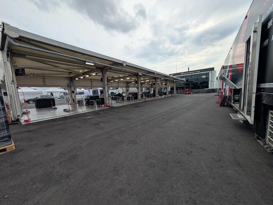 Rallitallin sisäpiha-alue service-parkissa. Oikealla reunalla näkyvissä rekka-autojen kyljet. Vasemmalla autojen huoltopilttuut, jossa ei vielä ole autoja. Keskellä tyhjää asvalttia.