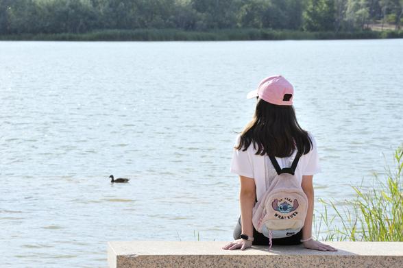 una adolescente con gorra, sentada en un banco de hormigón, observa unos patos (aunque en la foto solo sale uno) nadando en un estanque.