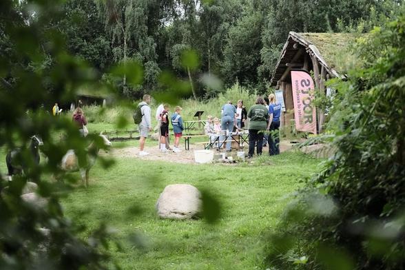 Durch eine Laubwand hindurch ist die Rekonstruktion eines steinzeitlichen Hauses zu sehen. Davor stehen mehrere Menschen, Tische, Bänke und eine Beachflag.