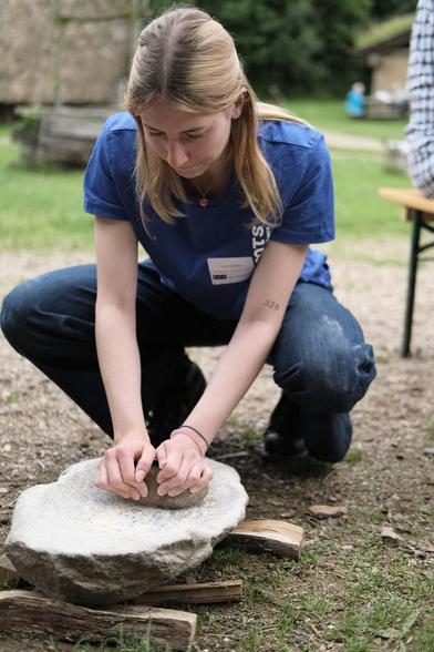 Eine Frau hockt vor einem etwa 40 Zentimeter langen und 30 Zentimeter breiten Stein mit einer flachen Mulde. In der Mulde liegen Gereidekörner. Die Frau bewegt einen kleinen runden Stein über die Körner.