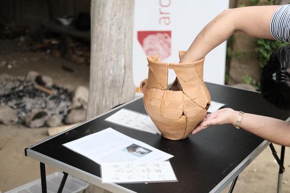 Ein rotbrauner Topf steht auf einem Tisch vor einem Holzpfosten. Eine Person, von der nur die Hände und Arme zu sehen sind arbeitet an dem Topf, der aus etlichen einzelnen Scherben besteht. Einige Scherben fehlen noch.
