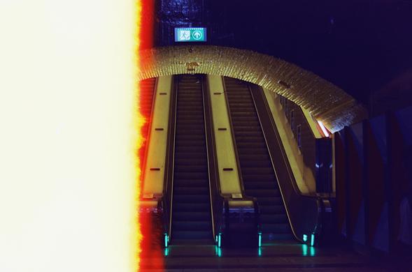 Analog photograph of the escalators and underground hallway at Stortinget metro station in Oslo. The left third of the photo is burnt out because this was the first (incomplete) frame on the roll of film. The other two thirds are dark with a blue tint due to the light leak, but it doesn't help that the hallway was also dark and the escalators crowned by a yellow ceiling. Toward the right edge, you can see the characteristic geometric wall panelling, unique to Stortinget station.