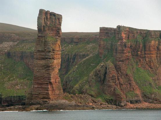The Old Man of Hoy. The image shows a view from the sea of a tall reddish stone rock stack towards the left of the frame. Behind it and continuing to the right of it are high cliffs partly covered in vegetation. The scene is in low sunlight coming from the right.
