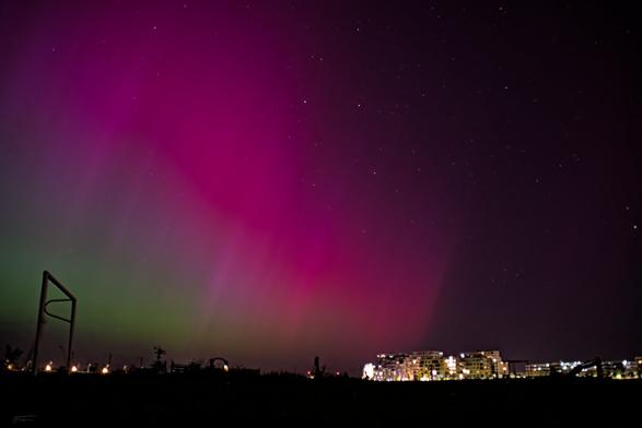 Das Bild zeigt eine nächtliche Szene mit einem beeindruckenden Himmel, der von einem leuchtenden Phänomen dominiert wird, das als Aurora oder Polarlicht bekannt ist. Die Aurora erscheint in verschiedenen Farben, darunter Grün und Violett, und erstreckt sich über den Himmel. Im Vordergrund sieht man eine dunkle Silhouette von Bäumen und Gebäuden, darunter auch einige Lichter, die auf eine Stadt oder ein Dorf hinweisen. Es gibt auch ein paar Strukturen, die wie Spielplatzgeräte oder ähnliche Konstruktionen aussehen. Der Himmel ist klar, und man kann einige Sterne sehen.
