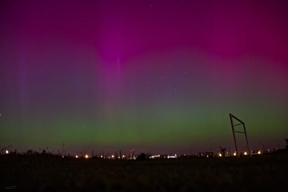 Das Bild zeigt eine nächtliche Szene mit einem beeindruckenden Himmel, der von einem leuchtenden Phänomen dominiert wird, das als Aurora oder Polarlicht bekannt ist. Die Aurora erscheint in verschiedenen Farben, darunter Grün und Violett, und erstreckt sich über den Himmel. Im Vordergrund sieht man eine dunkle Silhouette von Bäumen und Gebäuden, darunter auch einige Lichter, die auf eine Stadt oder ein Dorf hinweisen. Es gibt auch ein paar Strukturen, die wie Spielplatzgeräte oder ähnliche Konstruktionen aussehen. Der Himmel ist klar, und man kann einige Sterne sehen.