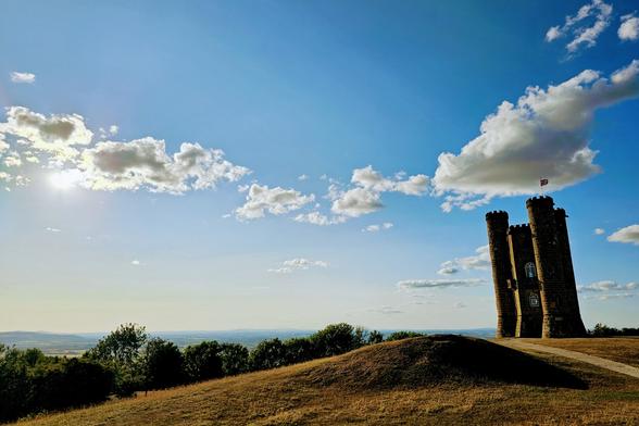A wide-angle, eye-level shot captures Broadway Tower, a tall, dark stone folly with turrets, standing prominently on a grassy hill under a vibrant blue sky dotted with fluffy white clouds. The sun, a bright starburst, is visible on the left side of the frame, casting a warm glow over the scene. To the left of the tower, the dark silhouettes of trees line the horizon, with a hazy, light blue sky above them indicating distant hills. A dirt path curves up the grassy hill towards the tower, inviting exploration.