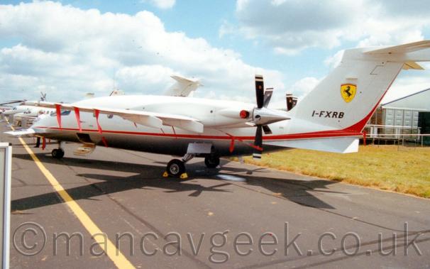 Rear side view of a twin propellor-engined BizProp with the engines mounted pointing backwards of the rear of the wings.
The plane is mostly white, with a grey belly, and a red stripe running along the body from the nose, before sweeping up into the rear of the tail.
The black registration "I-FXRB" is on the lower part of the tail, with a yellow shield in the middle, containing an image of a prancing black horse, with a thin red and green band at the top of the shield.
There are red covers on engine exhaust outlets, with red ribbons attached to several parts of the rear of the wings and on the fuselage.
The bottom half of the cabin door is hanging open, forming a set of stairs, while the upper half is closed.
There is a small aerodynamic lifting surface on the nose-cone.
Black tarmac taxiway fills the foreground, with grass in front of a white temporary building in the background on the right, with the forward fuselages of several BizJets in the background on the left.
Pale blue sky with large fluffs of white cloud fill the rest of the frame.