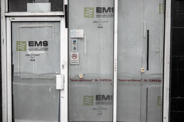 A glass-fronted shop has red text reading '"Great cuts forever" Mr. TOPPERS', behind the glass the view is blocked by a white sheet labelled 'EMS Construction'