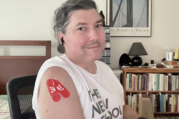 Selfie of a person sitting in an office chair in a New School T-shirt.  The sleeve of the T-shirt is raised to reveal two red bandages with the Walgreens logo, indicating vaccination sites.