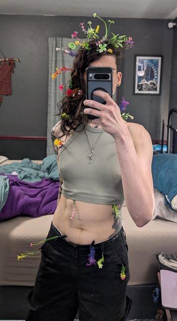 This enby taking a mirror selfie, thighs and up. They are wearing a sage green tank top, that can barely contains the nips. They're also wearing their salamander pendant and black cargo pants with spacious pockets. Their arm tattoo is slightly visible but obscured behind their torso. You can see some of their torso tattoo on the other side, with the mushrooms and wildflowers merging with the trail of sprouts growing from various nooks on their body.

There are too many to enumerate, but there is clearly a concentration of them at the top part of the head amidst the hair. Some have googly eyes, and there are a couple of glow-in-the-dark ducks as well. The mushroom one has a bowtie and a googly eye, and a few other sprouting plants have googly eyes as well. Some flowers, some vines, even a pink and blue butterfly. They continue down the hair, and also emerge from under the crop top. All along the shoulder straps and near the edge of the breasts. Various colors are represented: red and yellow, green and white, pink and white, bright deep red and green, blue and purple, etc. A pink and green viney one emerges from under the crop top traveling downwards parallel to the soft "gutters" formed by their abdominals. 

A black and white boyshorts waistband peeks out from behind the cargo pants as do yet even more sprouts. The color scheme here is more limited with a lot of green, pink, blue and some white and yellow.