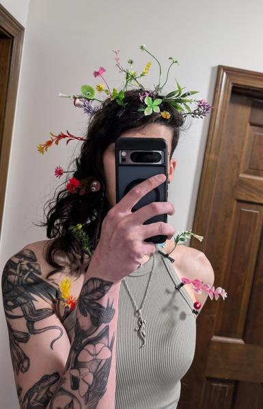 This enby taking a mirror selfie of their upper body. They are wearing a sage green tank top, and despite the ribbed texture, it barely contains the nipples. Their arm tattoos are in sharp definition. The potion bottle nestled among the leaves at the wrist, the owl with the portal and the two flying insectoids fluttering out. The toad by the bicep, looking at you with a smile, the Ichorite crystals and Eldritch tentacles emerging from the portals in the ravens wings at the shoulder with some mushrooms escaping up top. All visible with good detail. 

The most notable things, however, are all the different blooming sprouts. Too many to enumerate, but most of them are in the top part of the head amongst the hair. Some have googly eyes, and there are a couple of glow-in-the-dark ducks as well. The mushroom one has a bowtie and a googly eye, and a few other sprouting plants have googly eyes as well. Some flowers, some vines, even a pink and blue butterfly. They continue down the hair, and also emerge from under the crop top. All along the shoulder straps and near the edge of the breasts. Various colors are represented: red and yellow, green and white, pink and white, bright deep red and green, blue and purple, etc. Near one of the breasts is a red sphere with its own single googly eye as well, staring directly at the viewer.