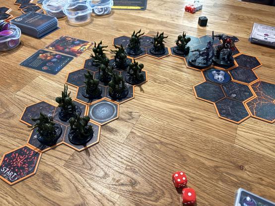 Das Bild zeigt eine Szene aus einem Brettspiel auf einem Holztisch. Im Vordergrund sind mehrere sechseckige Spielplatten mit orangefarbenen Rändern angeordnet, die eine Karte oder ein Spielbrett darstellen. Auf diesen Platten stehen kleine, dunkelgrüne Figurinen, die Monster oder Kreaturen darstellen, sowie größere, dunkelgrau-braune Figuren, die wahrscheinlich Spielercharaktere sind. Einige der Kreaturen sind in einer Reihe angeordnet, während andere sich in einer Gruppe befinden.

Im oberen Bereich des Bildes sind mehrere Karten und Spielzubehör sichtbar, darunter eine Karte mit der Aufschrift "START" und mehrere Karten mit verschiedenen Symbolen und Texten. Einige transparente Behälter enthalten weitere Spielteile. Zwei rote Würfel liegen auf dem Tisch, und ein schwarzer Würfel ist ebenfalls sichtbar.

Die Szene vermittelt den Eindruck eines aktiven Brettspiels, bei dem die Spieler strategisch positionieren und bewegen, um ihre Figuren zu bewegen und zu interagieren.
