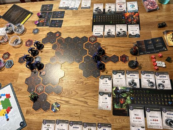 Das Bild zeigt eine ansprechende Szene eines Brettspiels auf einem Holztisch. Im Zentrum des Spiels befindet sich ein hexagonales Feld, das aus verschiedenen Feldern besteht, die wahrscheinlich verschiedene Zonen oder Räume im Spiel darstellen. Auf diesem Feld sind mehrere kleine Figuren in dunklen Farben platziert, die wahrscheinlich Spielerfiguren sind.

Um das Spielfeld herum sind verschiedene Spielkomponenten verteilt. Links oben sind mehrere Karten und Zettel zu sehen, die wahrscheinlich Spielregeln oder Spielzüge enthalten. Rechts oben befinden sich mehrere Karten, die wahrscheinlich verschiedene Spielkarten oder Fähigkeiten darstellen. Einige dieser Karten haben Zahlen und Symbole, die wahrscheinlich die Spielmechanik beeinflussen.

Unter den Karten befindet sich ein Spielbrett mit verschiedenen Zahlen und Symbole, das wahrscheinlich die Spielzüge oder die Spielstatus verfolgt. Rechts neben dem Brett sind mehrere Würfel und weitere Spielkomponenten wie Steine und Figuren zu sehen.

Im unteren Bereich des Bildes sind mehrere Karten mit Namen wie "Death Glaive", "Nature Shot" und "Strike of Gort" sichtbar, die wahrscheinlich verschiedene Spielkarten oder Fähigkeiten darstellen. Links unten ist ein kleines Kartenblatt mit einer Karte, die wahrscheinlich eine Karte aus dem Spiel ist.

Die gesamte Szene ist gut organisiert und zeigt eine ansprechende und komplexe Brettspielumgebung.