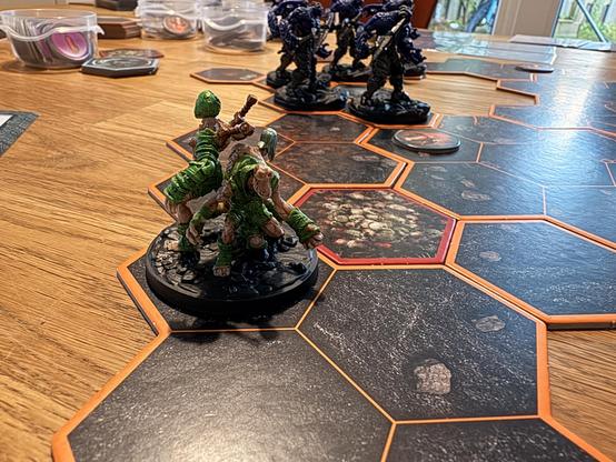 Das Bild zeigt eine Szene aus einem Brettspiel auf einem Holztisch. Im Vordergrund befinden sich mehrere miniaturisierte Figuren, die auf hexagonalen Spielplatten mit orangefarbenen Rändern platziert sind. Die Figuren im Vordergrund sind grün und scheinen aus einer natürlichen Umgebung zu stammen, mit einer Figur, die einen Speer hält. Im Hintergrund sind weitere Figuren in dunkelblauer Farbe zu sehen, die eine militärische Erscheinung haben. Die Spielplatten haben verschiedene Muster und Farben, darunter ein hexagonales Feld mit einem roten Rand, das möglicherweise eine besondere Bedeutung hat. Im Hintergrund sind auch einige Spielkarten und Behälter mit Spielteilen zu sehen. Die Szene vermittelt den Eindruck eines aktiven Brettspiels, bei dem die Figuren strategisch positioniert sind.