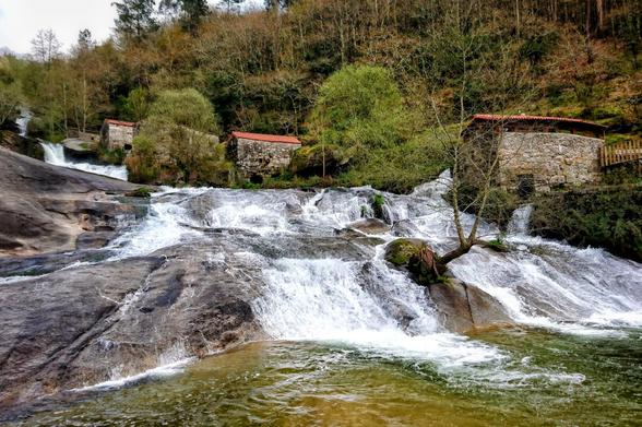 A wide shot of a multi-tiered waterfall cascading over dark grey rocks into a river. On the right bank, nestled amongst the trees, are several old stone buildings with reddish-brown tiled roofs. The water is white and frothy where it rushes over the rocks, and a clear, dark green in the calmer pools. Lush green and brown trees cover the hills in the background under a soft, overcast sky.