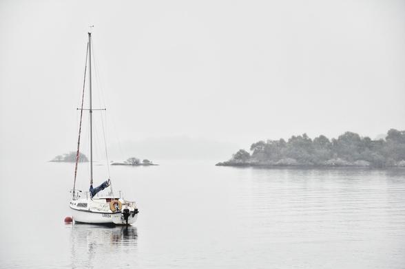 Segelboot auf ruhigem Wasser nahe bewaldeter Insel unter nebligem Himmel