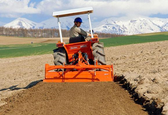 A small orange-red tractor is pulling a rotavator behind it. The tractor is on a slope and tillting left along the roll axis. The rotavator attachment is however level with the horizon and the resulting smooth kultivated soil is level behind it, despite the tractor bouncing over a lumpy plowed field. There's an edge of raised soil on the left (downhill side) and a cut into the soil on the right (uphill) side.