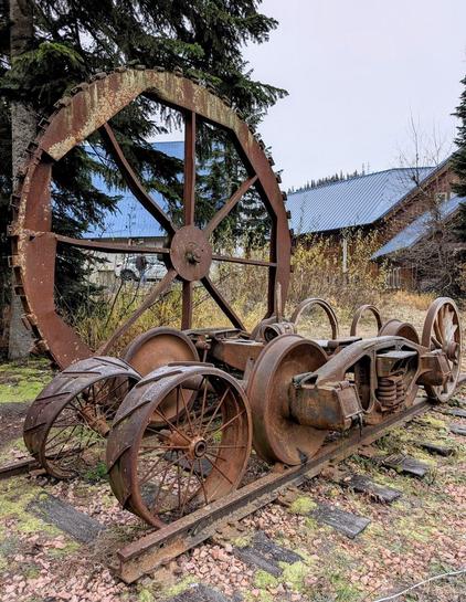 An old, rusted set of train parts sits on abandoned narrow tracks at Snoqualmie Pass, WA. The structure includes a massive spoked wheel with gear-like teeth around its rim, connected to several smaller metal train wheels and a heavy steel frame with exposed springs and axles. Moss and vegetation grow through the gravel beneath, showing years of weathering. Behind the relic, evergreen trees and buildings with bright blue metal roofs stand under an overcast sky, giving the scene a sense of history and abandonment.