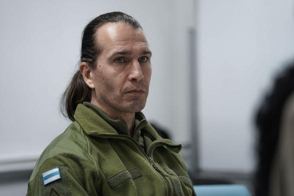 Caesar in a green military jacket with an Free Russia flag patch sits against a blurred background, appearing thoughtful.