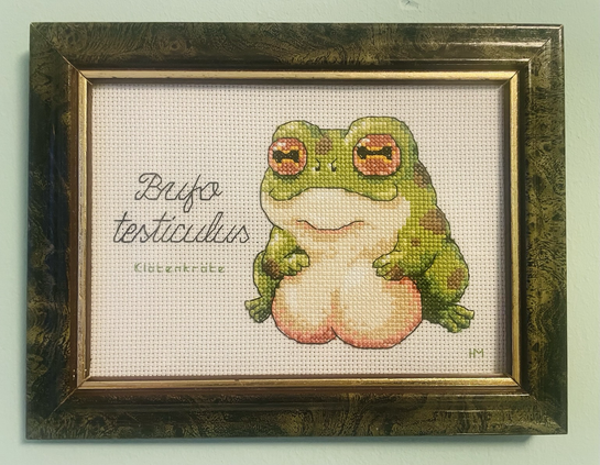 In einem grüngoldenen Bilderrahmen ist eine dicke grünbraune Kröte mit enormen Hoden gestickt. Ihre kleinen Patscherhändchen ruhen entspannt auf den Testikeln. Daneben steht „Bufo testiculus - Klötenkröte“