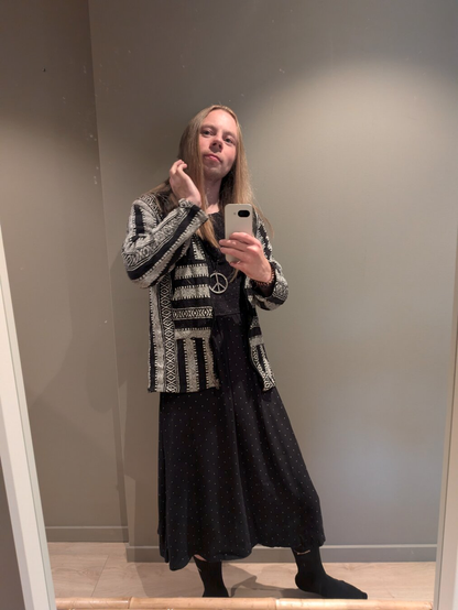 A gender-fluid person takes a mirror selfie with a smartphone, wearing a polka dot dress and a black and white jacket, along with a peace necklace. The plain wall in the background provides a simple, minimalist backdrop.
