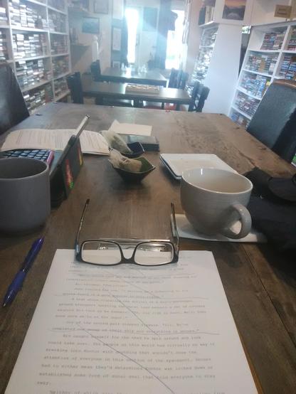 From the bottom up. Clipboard holding printed pagss of my manuscript. Two cups of tea, on for me on the right and one for my daughter on the left. Teabag bowls and treat plates. End of our table then two more tables presently unoccupied. Front door too to the left and window to the right. On both sides of this line are shelves filled with used books.