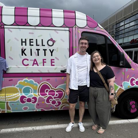 A couple standing in front of a large van that is decked out in pink and various Hello Kitty icons and pastries in a similar 2D outline art style. Next to the couple, the van reads “Hello Kitty Cafe”. A man is wearing an open white button up shirt sleeve shirt with black shorts and a woman is wearing a black v-neck shirt with greyish green pants.