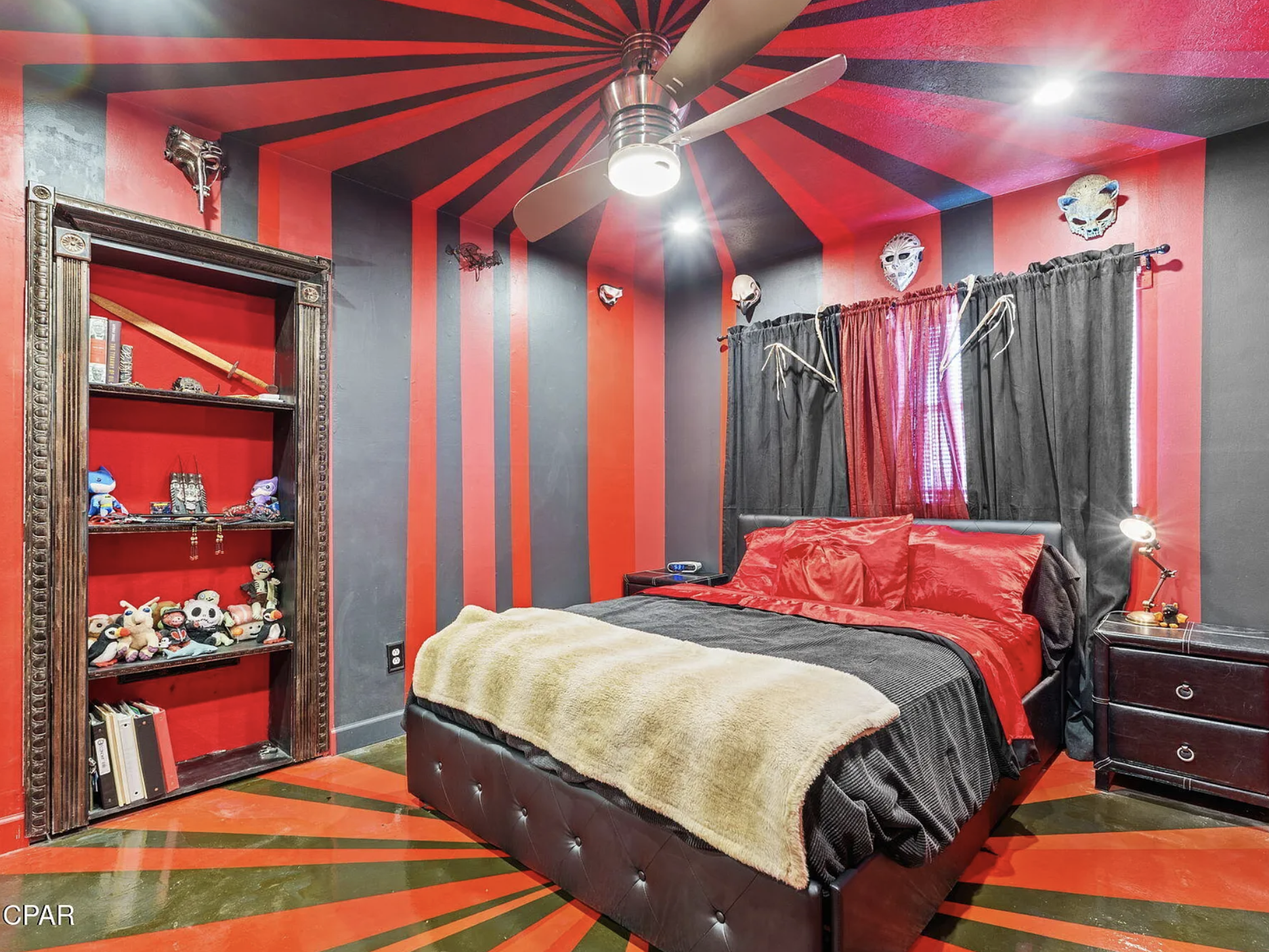 A bedroom with black and red painted stripes up the walls and across the ceiling and the same stripes are on the floor. It kind of  of looks like a goth circus tent. There are also creepy masks above the bed.