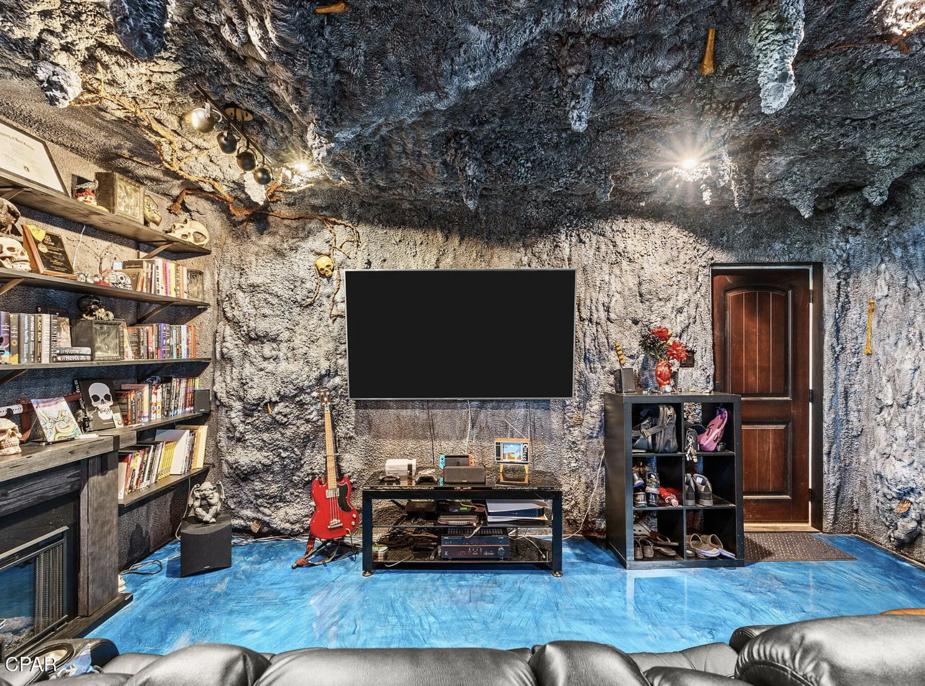 A room made to look like a cave, despite it not being a cave. The walls and ceiling are grey and very lumpy, with bits hanging down from the ceiling. There is also a tv, guitar, and a blue rug.