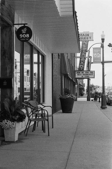 A black and white film photo of a quiet small-town sidewalk in Detroit Lakes, Minnesota. The image shows storefronts, including a coffee shop and a diner with a vintage hanging sign reading “Main Street Restaurant.” Empty chairs and flower planters line the sidewalk, evoking a still, almost frozen-in-time atmosphere.