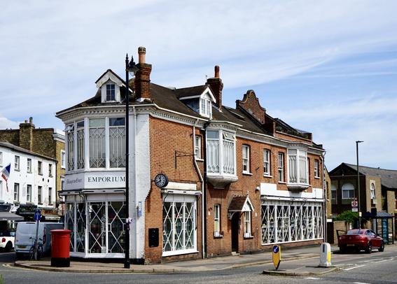 Vue en biais d’un bâtiment sur deux niveaux en ville, style architecture victorienne, à l’angle de deux rues. D’un côté, la façade est en brique orange, égayée par deux logettes blanches et par des croisillons blancs protégeant des vitrines en verre au rez de chaussée. Il y a une petite horloge rétro à l’angle. De l’autre côté, on ne voit que le début de la façade, elle semble occupée par des baies vitrées à l’étage et à nouveau des vitrines à croisillons en bas. L’enseigne sur le côté dit « Hampton Court Emporium ». Il y a encore une boîte aux lettres rutilante devant l’angle pour parachever cette vision toute anglaise.
