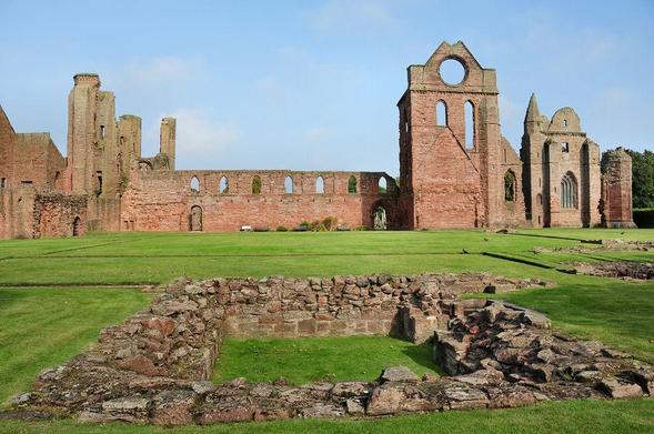 Arbroath Abbey. The image shows a low pinkish stone square foundation in the foreground, set amongst grass with other low foundations stretching back from it. In the distance are the imposing ruins of the abbey, rising to considerable height and made of pinkish stone. The sky is blue.