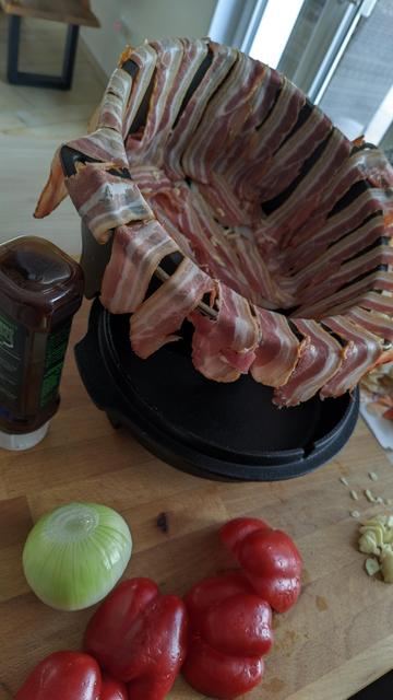 ein Dutch Oven steht auf einem Holzbrett, eine Schicht Zwiebeln und Speck ist schon eingelegt, drumherum noch ein paar andere Zutaten