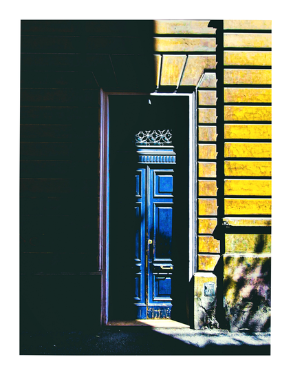 The photo is of a dark blue door set within a building's facade.  The building's exterior is divided into two contrasting sections; one side is dark and shadowy, while the other is brightly lit and has a yellowish-gold tone. There's a ray of sunlight which illuminates the door and a small portion of the ground in front of it. The door has ornate iron wrought detailing in the top.