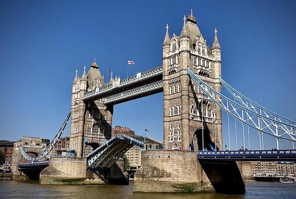 Tower Bridge in London, featuring two prominent towers with pointed roofs connected by upper walkways, set against a clear blue sky. The bridge is partly raised, displaying its bascule (drawbridge) mechanism in action. Visible are intricate architectural details on the Victorian Gothic towers and the blue and white color scheme of the bridge structure. A UK flag flutters atop one tower. The Thames River and riverbanks are visible, with architectural buildings in the background.