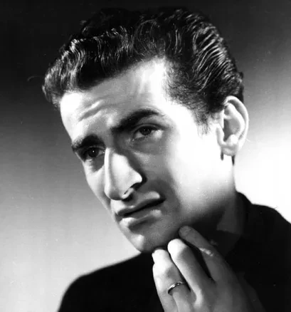 Photographie en noir et blanc de style portrait harcourt d'un homme plutôt jeune  peau claire, cheveux noirs épais et gominé, l'air soucieux, une mains sous le menton.  Il est vêtu d’un costume sombre avec une chemise blanche visible au niveau du col. L’arrière-plan est uniforme et gris clair.