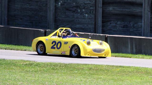 Photograph of a yellow “bug-eye” Austin-Healey Sprite seen from the side traveling to the right on a racetrack with grass in front and a retaining wall behind. The headlights and grill form its classic smile.