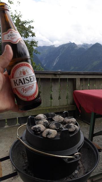 ein Dopf steht in einem Kugelgrill, darauf glühende Briketts, jemand hält eine Flasche Bier ins Bild und im Hintergrund sind Berge zu sehen