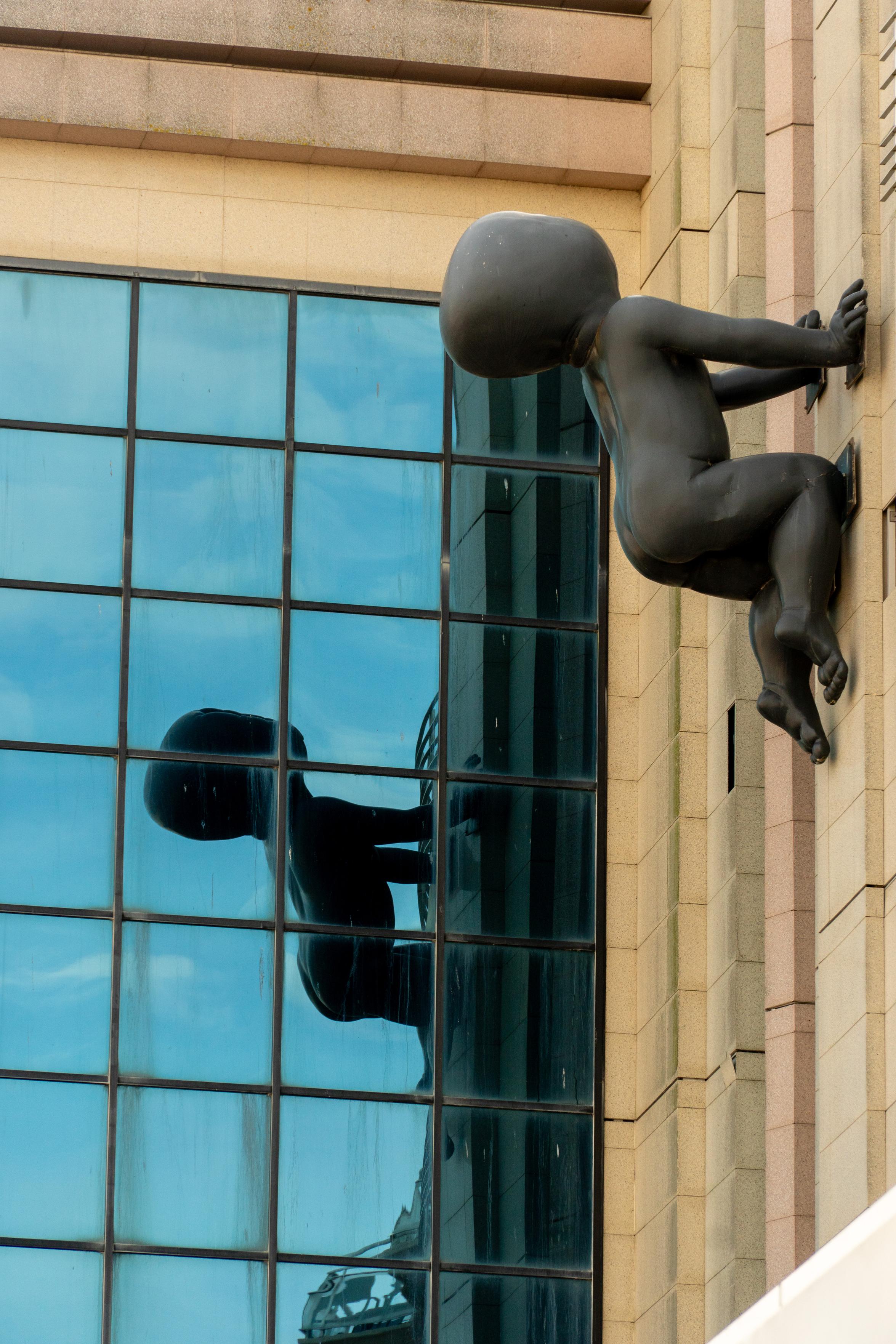 A stone baby climbing up the side of a building, reflected in windows of the same building.