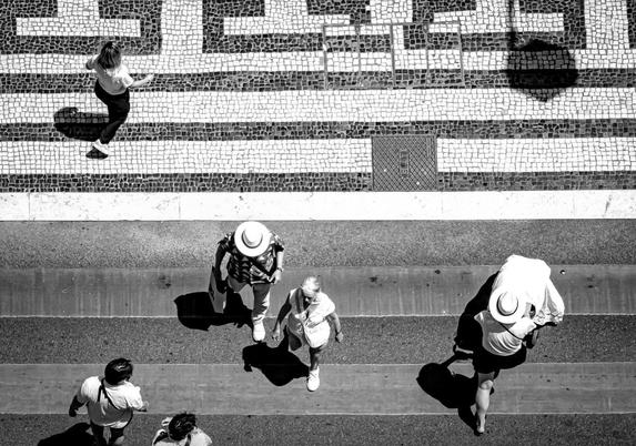 Aerial view of a black and white scene with several people walking on cobblestone and paved pathways. Geometric tile patterns and shadows are visible on the ground. The individuals cast distinct shadows, and one person carries a large white bag or item. Two people wear wide-brimmed hats, and all figures are oriented in various directions.