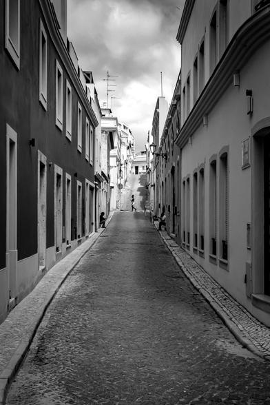 Narrow cobblestone street flanked by tall buildings with multiple windows, leading to an inclined pathway. A few people are sitting along the side and one person is walking near the top of the incline. Cloudy sky overhead adds to the contrast in the black and white photograph.