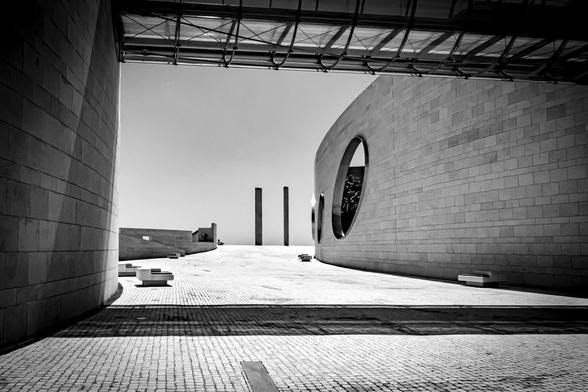 Modern architectural scene in black and white with geometric structures. Foreground features a tiled plaza bordered by curved stone buildings. Large round windows are visible on the right building. Overhead, a glass and metal structure creates shadow patterns on the ground. Two tall, slender pillars rise in the distance under a clear sky.