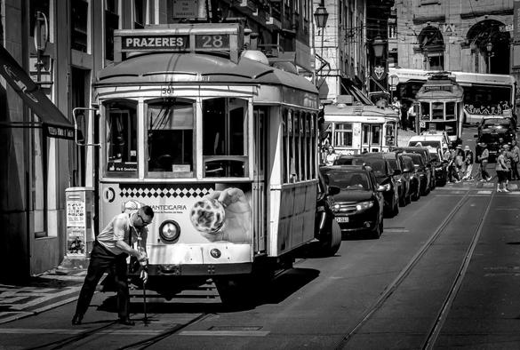 Historic tram labeled "28" on a city street with tracks, parked cars, and a pedestrian crossing in front. The tram features an advertisement with a hand holding a pastry. More trams and people visible in the background, buildings and wires overhead. Image in black and white.
