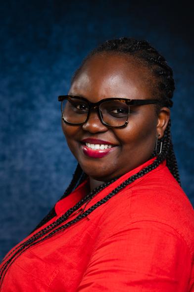 A person with braided hair wearing glasses and a red shirt is smiling warmly. The background is a deep blue, creating a striking contrast with the red shirt, drawing attention to the person’s joyful expression.