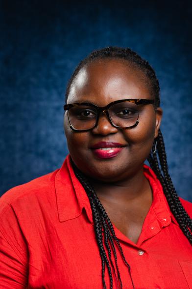 A person with braided hair and glasses is smiling. They are wearing a bright red shirt, and the background is a textured deep blue, creating a vibrant contrast.