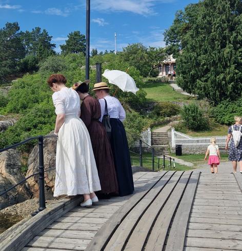 Three women in historical dresses stand on a wooden bridge, looking over the railing toward the water. One holds a white parasol, and all wear 1905-inspired clothing. It's a sunny day at a heritage site with green trees, grass, and an old building in the background. A small child in a pink dress walks away on the right.