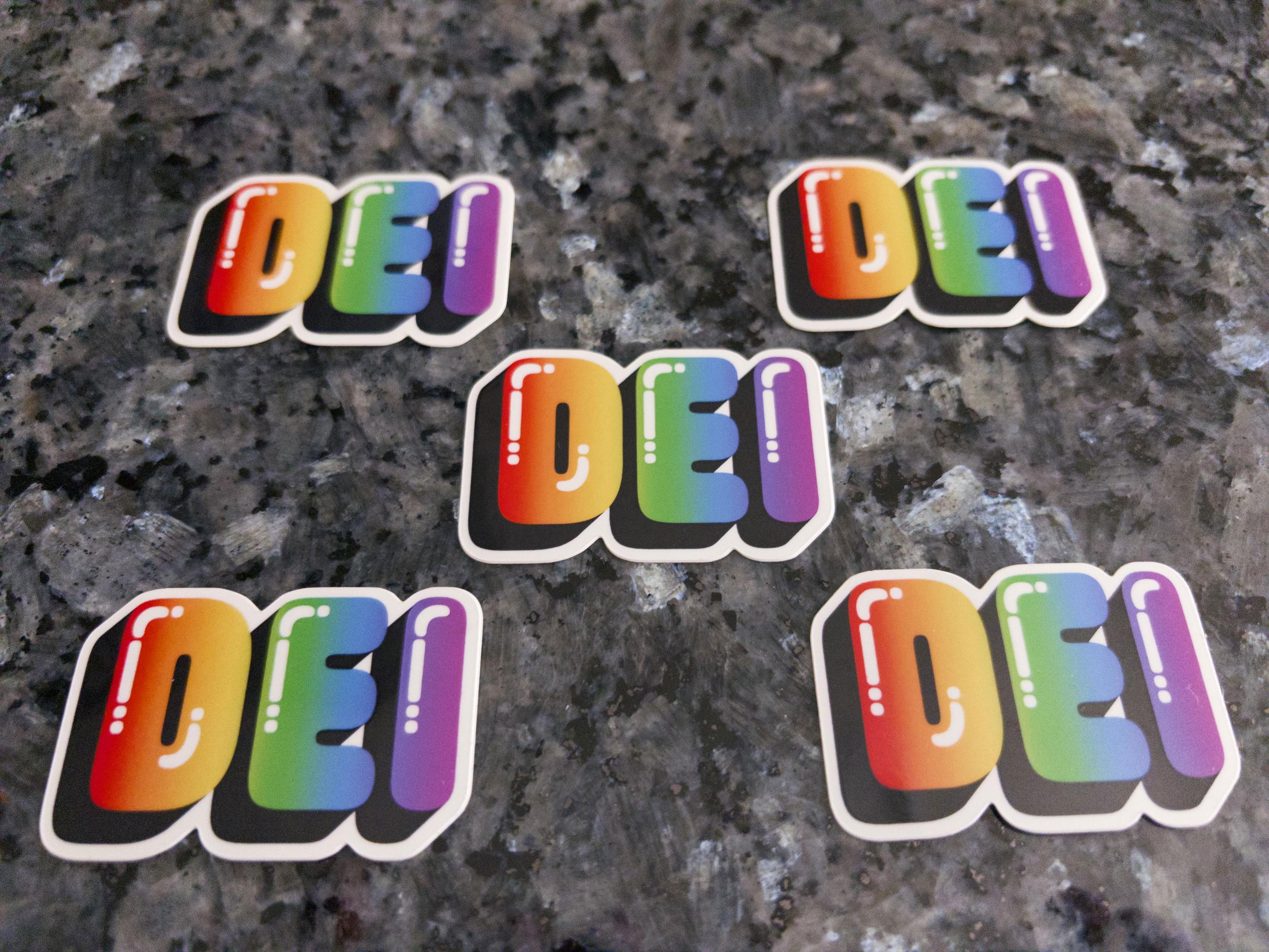 5 stickers featuring the letters DEI in a rainbow spectrum with a shine effect, positioned on a dark granite countertop.