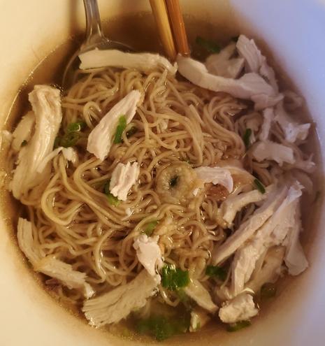 Close-up overhead shot of a bowl of chicken noodle soup with broth, egg noodles, and shredded chicken, garnished with green onions, with a spoon and chopsticks in the bowl.